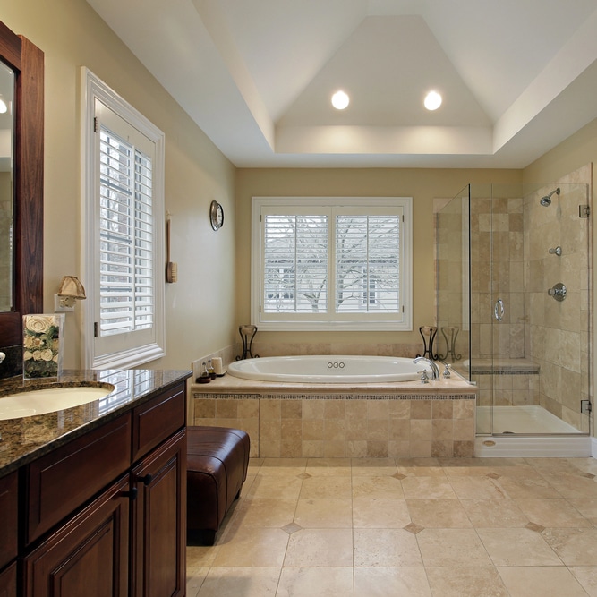 Image of a beautiful modern bathroom with subtle earthtone tiles, a dark oak wood double vanity, a glass enclosed shower, and a spa like tub with a window.