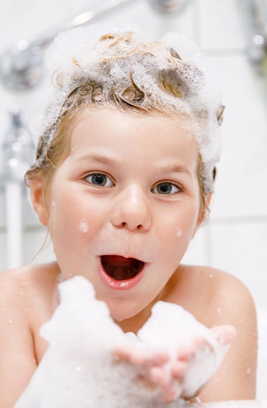 Cute image of a child in a bubble bath covered in bubbles with a happy but mischievous look about to blow a hand full of bubbles at the camera.