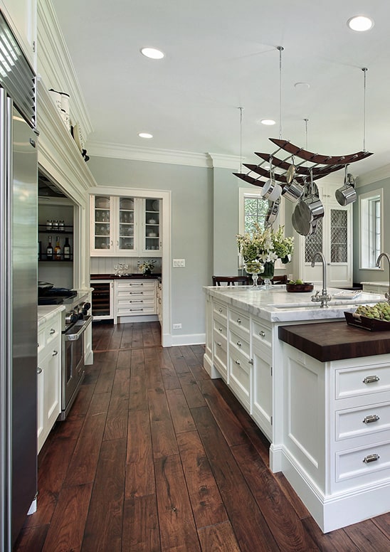 Image of a newly renovated kitchen with modern appliances and colonial style white cabinates with glass wooden grills separating rectngular panels of glass and a beautiful white kitchen island with stainless steel pots hanging overhead by ceiling hooks.