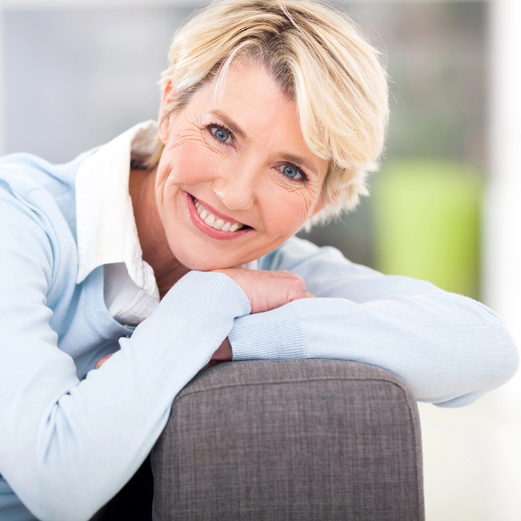 Lovely blond-haired middle-aged woman dressed in a blue blouse and smiling with her head resting on her arms that are folded over the arm of a couch.